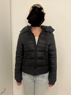 Svart pufferjacka med huva från Gina Tricot - Svart pufferjacka från Gina Tricot med avtagbar huva och två snedställda fickor med dragkedja. Jackan har quiltad design, hel dragkedja framtill och elastiska muddar vid ärmslut. Perfekt för kalla dagar och har en snygg, lite kortare passform. Skriv om du har någon fråga ❤️