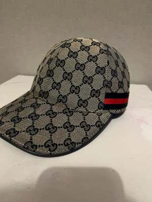 Gucci keps med GG-mönster och röd detalj - Snygg beige och svart keps från Gucci med klassiskt GG-mönster över hela kepsen. Har en röd och svart rand på sidan och justerbar rem bak. Materialet är bomull och polyester, perfekt för dig som vill sticka ut med en lyxig streetstyle.
