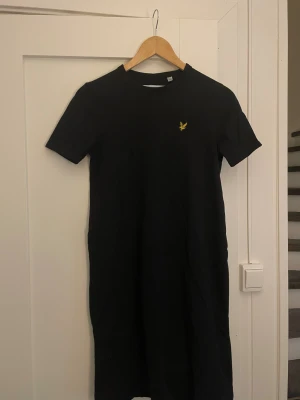 Svart t-shirtklänning från Lyle & Scott - Svart t-shirtklänning från Lyle & Scott i 100% ekologisk bomull. Klassisk rund hals, korta ärmar och gul broderad logga på bröstet. Enkel och stilren design som är lätt att matcha med sneakers eller boots.