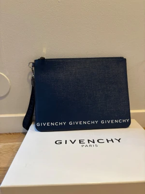 Mörkblå kuvertväska från Givenchy - Snygg mörkblå kuvertväska från Givenchy med vit logotyptext längs nederkanten. Väskan har en avtagbar handledsrem med Givenchy-tryck och stängs med dragkedja. Materialet är strukturerat skinn som ger en exklusiv känsla. Perfekt för att bära dina essentials. 30 x 23cm                                         Kartong etc ingår.   Skickas inom 24h. Priset är inte hugget i sten.  