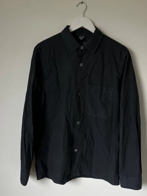 Svart overshirt från Arket - Svart/mörkgrå overshirt från Arket i bomull. Klassisk krage, knappstängning framtill och en bröstficka. Långärmad och med en clean, minimalistisk look som funkar till många stilar. Perfekt för dig som gillar enkel och stilren design. 5L9