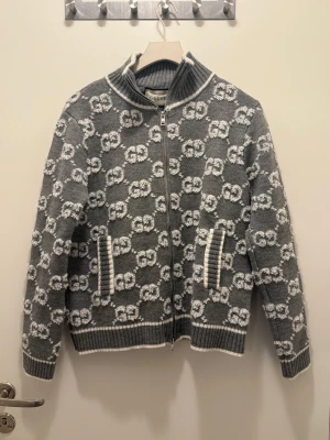 Grå stickad kofta från Gucci - Säljer en grå och vit stickad kofta från Gucci med GG-mönster över hela plagget. Koftan har dragkedja framtill, ribbad krage, muddar och nederkant. Tillverkad i 100% ull och har två fickor med vit ribbad kant. Perfekt för dig som gillar lyxiga detaljer.