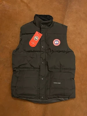Svart dunväst från Canada Goose - Svart dunväst från Canada Goose med klassisk logotyp på bröstet och tryckknappar framtill. Västen har två stora fickor och hög krage. Materialet är slitstark polyester och dunfyllning som håller värmen. Perfekt för kyliga dagar och har en stilren look.