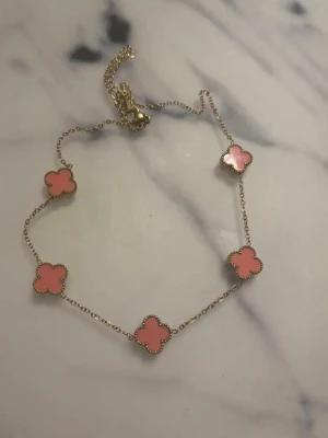 Guldfärgad halsband med rosa klöverdetaljer - Guldfärgat halsband med flera söta klöverformade berlocker i rosa nyans. Klassisk och feminin design som passar perfekt både till vardag och fest. Lätt att matcha med andra smycken eller bära som statement i sig själv.