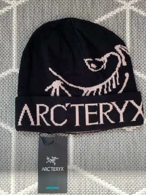 Svart Arc'teryx mössa, Aldrig Använt! - Snygg svart mössa från Arc'teryx med stor, stickad logga och fossilmotiv i ljusbeige. Mössan är tillverkad i mjukt stickat material och har en bred uppvikt kant. Perfekt för kalla dagar och outdoor-aktiviteter.