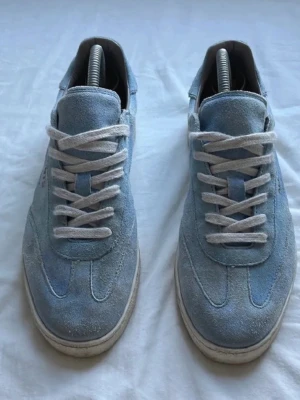 All Saints ljusblå sneakers mocka - Säljer ett par ljusblå sneakers från All Saints i mjuk mocka med vita snören och platt sula. Skorna har en rund tå och diskret guldfärgad logga på sidan. Perfekt för dig som gillar stilrena och fräscha sneakers med snygg retrovibe.