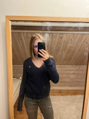 Marinblå stickad tröja från Ralph Lauren - Säljer en marinblå stickad tröja från Ralph Lauren med klassisk kabelstickning och v-ringad hals. Tröjan har långa ärmar och en vit broderad logga på bröstet. Perfekt för dig som gillar stilrena och tidlösa plagg.