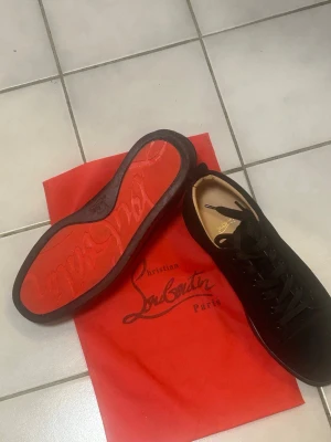 Svarta sneakers från Christian Louboutin - Säljer ett par svarta sneakers från Christian Louboutin med klassisk röd sula och snyggt guldfärgat logo-tryck på innersulan. Skorna har snörning och är tillverkade i mocka med rund tå och platt sula. Perfekt för dig som gillar exklusiva och stilrena sneakers.
