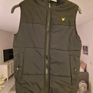 Svart dunväst från Lyle & Scott - Säljer en svart dunväst från Lyle & Scott med gul logga på bröstet. Västen har dragkedja framtill och två fickor med knappstängning. Perfekt att slänga över en hoodie för extra värme. Snygg och enkel design som funkar till många stilar.