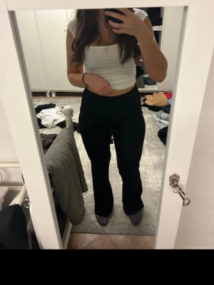 Svarta bootcut byxor med dragkedja - Säljer ett par svarta bootcut byxor med hög midja och smal passform upptill som blir vidare nedtill. Byxorna har en diskret dragkedja framtill och är gjorda i ett mjukt material som sitter skönt mot kroppen. Perfekta om du gillar stilrena och trendiga silhuetter.