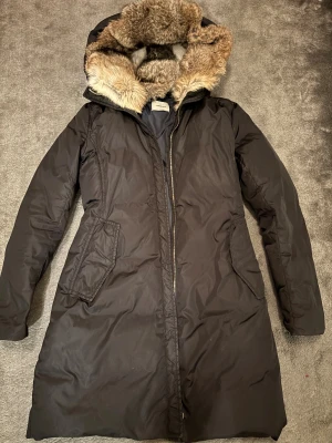 Svart dunjacka med päls från Lempelius - Lång svart dunjacka från Lempelius med stor huva och fluffig päls. Jackan har dragkedja, två snedställda fickor och en figurnära passform. Perfekt för kalla dagar när du vill vara både varm och snygg. 