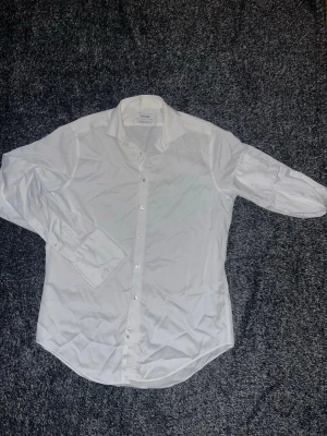 Vit slim fit skjorta från Calvin Klein - Säljer en stilren vit skjorta från Calvin Klein med slim fit passform och non-iron material. Skjortan har klassisk krage, långa ärmar och knappar framtill. Perfekt för dig som gillar en clean och modern look. Tveka inte på att höra av dig vid funderingar;) /Isak