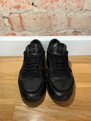 Svarta Valentino Garavani Rockrunner sneakers - Svarta Valentino Garavani Rockrunner sneakers med coola nitar på hälen, snörning och rund tå. Skorna har en mix av skinn och mocka i ovandelen, vilket ger en schysst kontrast. Snygg och stilren design med svarta detaljer och platt sula.