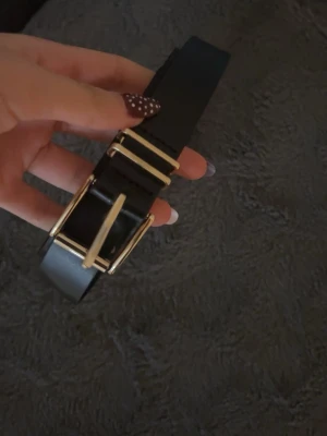 Svart skärp från H&M med gulddetaljer - Säljer ett svart skärp från H&M i storlek XS med snygga guldfärgade metallspännen. Skärpet har en stilren och klassisk design som passar till många olika outfits. Perfekt för att ge din look lite extra edge.