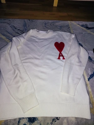 Vit stickad tröja med rött hjärta - Säljer en vit stickad tröja från AMI Paris med ett stort rött hjärta och bokstaven A broderat på bröstet. Tröjan har rund hals, ribbade muddar och är långärmad. Perfekt för dig som gillar stilrena detaljer och vill sticka ut med en ikonisk design.