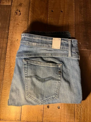 Replay ljusblå jeans - Säljer ett par ljusblå jeans från Replay med snygga slitningar och dekorativa nitar på bakfickorna. Jeansen har klassisk femficksdesign, normal passform och raka ben. Materialet är mjukt jeans-tyg i bomull och de har en tydlig Replay-logga vid fickan.