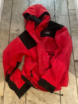 Röd Goretex jacka från The North Face - Säljer en röd och svart Goretex från The North Face med huva och justerbara muddar. Jackan har dragkedja och knappar framtill, samt snörning vid midjan. Perfekt för dig som gillar att vara ute och vill ha en cool och funktionell jacka.