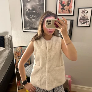 Vit pälsväst med fickor från Zara  - Säljer en supermjuk och fluffig vit pälsväst med två stora fickor framtill. Västen är ärmlös och har en rak passform som ger en clean och stilren look. Perfekt att slänga över en hoodie eller långärmad tröja för extra värme.