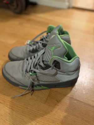 Nike Air Jordan 5 grå/grön sneakers - Säljer ett par Nike Air Jordan 5 sneakers i grått med gröna detaljer och insida. Skorna har snörning, hög modell och platt sula. Materialet är skinn och syntet med meshpaneler på sidorna. Jumpman-logga på plös och häl. Perfekt för dig som gillar streetwear.