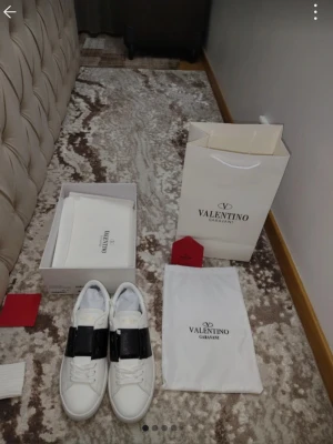 Valentino Garavani vita sneakers - Säljer ett par snygga vita Valentino Garavani sneakers med bred svart rem över foten. Skorna har rund tå, platt sula och klassisk snörning. Kommer med originalkartong, dustbag och shoppingpåse. Perfekta för dig som gillar stilrena designer.