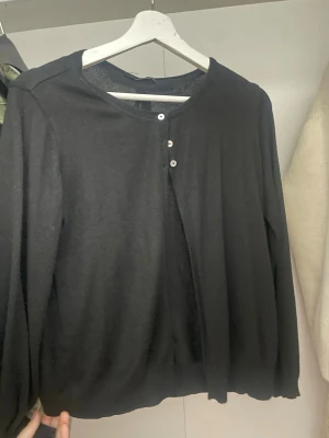 Svart kofta med knyt och knappar - Svart tunn kofta med rund hals och tre små knappar framtill. Baktill finns en snygg knytning som ger extra detalj. Långärmad och tillverkad i mjukt stickat material, perfekt för lager-på-lager under höst och vinter.