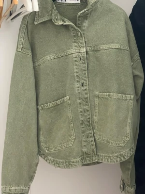 Grön jeansjacka från Zara - Säljer en grön jeansjacka från Zara med två stora fickor framtill och klassisk krage. Jackan har knappar hela vägen och är i en loose passform, perfekt att slänga på över en hoodie. Materialet är robust denim och färgen är en snygg, lite urtvättad grön.