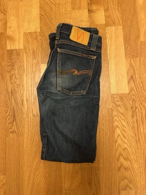 Nudie jeans slim fit  - Snygga mörkblå jeans från Nudie Jeans med klassisk femficksdesign och orangea sömmar. Jeansen har rak passform och är tillverkade i slitstarkt denimtyg av bomull. Bakfickorna har Nudie Jeans signaturbroderi och en läderpatch med logga i midjan. 36 cm i midjan och 71 på längden 