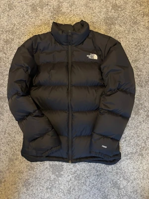 Svart dunjacka från The North Face - Säljer en svart pufferjacka från The North Face med 700-fill power dun. Jackan har hög krage, dragkedja framtill och logga på bröstet och ryggen. Perfekt för kalla dagar och har en clean, stilren look som funkar till det mesta.