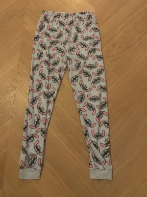 Ljus blå mjukisbyxor med Stitch-mönster - Supermysiga grå mjukisbyxor från Primark med Stitch-motiv över hela byxan. Byxorna har elastisk midja och är gjorda i mjukt fleece-material. Perfekta för chill hemma och har coolt tryck med rosa detaljer på öronen.