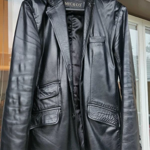 Svart skinnkavaj från MEC & CO - Svart kavaj i äkta skinn från MEC & CO. Unik design med 4 framfickor och snyggt foder på insidan. Jackan har krage och knäppning framtill, perfekt för dig som vill ha en stilren och edgy look. Vintage plagg! Det högsta kvalitet av leder som inte produceras längre! Unik! Köpt för 30 år sedan i Italien.Kavajen är som ny, men på grund av att skinnet  på baksidan under nacken har losnat lite1 cm( ej djupt) så man kan applicera Leather Filler, om man vill( se sista bilden). Kavajen är tung och varm! 
