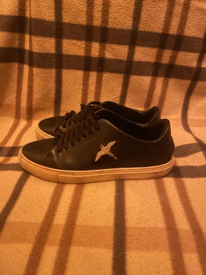 Svarta sneakers från Axel Arigato - Svarta sneakers från Axel Arigato i skinn med vit sula och broderad fågel på sidan. Klassisk låg modell med snörning och rund tå. Snygg minimalistisk design och diskret logga på sidan. Perfekta för dig som gillar stilrena och trendiga skor.
