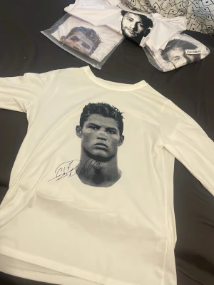 Vit långärmad topp med porträtttryck - Säljer en vit långärmad topp med ett stort svartvitt porträtttryck framtill och en tryckt autograf under motivet. Toppen har rund halsringning och är gjord i ett mjukt, slätt material som känns skönt mot huden. Perfekt för dig som gillar unika prints och vill sticka ut.