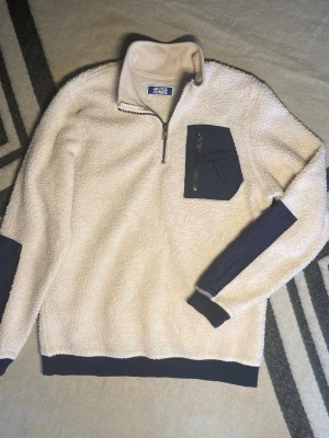 Jack & Jones Fleece - Mysig Fleece i beige från Jack & Jones med mörkblå detaljer på ficka, ärmslut och nederkant. Jackan har en hög krage, half zip och en praktisk bröstficka med dragkedja. Perfekt för dig som gillar chill och stilren look.