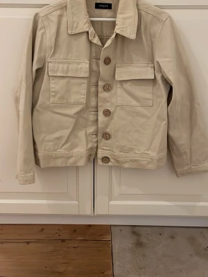 Beige jeansjacka från Pieces - Snygg beige jeansjacka från Pieces i storlek M. Jackan har klassisk krage, stora bröstfickor med lock och knappar framtill. Materialet är robust bomull och passformen är normal. Perfekt att slänga på över en hoodie eller t-shirt.