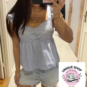 Blårandig blus med volangaxlar - Supersöt blus med blå och vita ränder, volangdetaljer på axlarna och lätt v-ringad hals. Blusen har en luftig passform och är perfekt för varma dagar. Materialet känns som bomull och toppen är ärmlös med breda axelband.