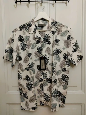 Vit blommig skjorta Mel Richman M - Säljer en helt ny vit kortärmad skjorta från Mel Richman i storlek M. Skjortan har ett snyggt blommigt mönster i svart, grått och grönt, klassisk krage och knäppning framtill. Perfekt för sommarens alla äventyr.