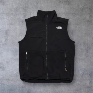Svart fleeceväst från The North Face - Svart fleeceväst från The North Face med hög krage och hel dragkedja framtill. Västen har två praktiska fickor med dragkedja och en bröstficka. Loggan är broderad i vitt på både fram- och baksidan. Perfekt för lager-på-lager och outdoor.