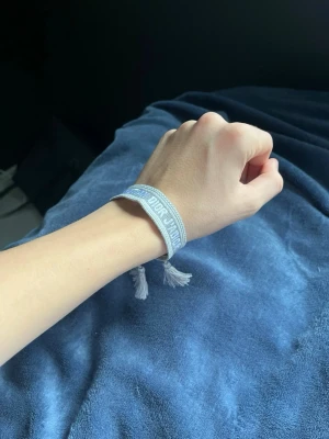 Grå Christian Dior J'Adior armband - Snyggt grått armband från Christian Dior med texten 'J'Adior' broderad i vitt. Armbandet är gjort i bomull och har dekorativa tofsar i ändarna. Perfekt accessoar för att lyfta din outfit med lite lyxig vibe.