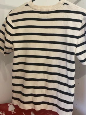 Randig vit t-shirt med svarta ränder - Snygg vit t-shirt med svarta horisontella ränder och rund hals. T-shirten har korta ärmar och är gjord i ett mjukt bomullsmaterial som känns skönt mot huden. Perfekt för dig som gillar klassisk och enkel stil med en twist.