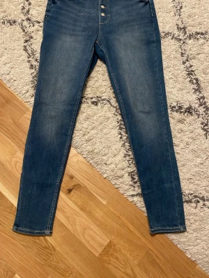 Blå skinny jeans från C&A, strl 42 - Säljer ett par blå skinny jeans från C&A i storlek 42. Jeansen har klassisk femficksdesign, knappgylf med fyra knappar och smal passform hela vägen ner. Materialet är stretchigt jeans och färgen är mörkblå med lätt tvättade detaljer.