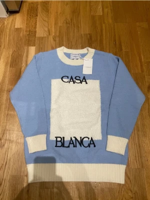 Blå stickad tröja Casablanca S - Säljer en helt ny ljusblå stickad tröja från Casablanca med kontrasterande vita ribbade muddar och krage. Framsidan har en stor vit fyrkant med broderad text 'CASA BLANCA' i svart. Tröjan är gjord i bomull och har långärmad passform.