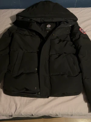 Canada Goose MacMillan - Säljer en svart pufferjacka från Canada Goose i storlek M. Jackan har huva, dragkedja och knappar framtill samt flera fickor. Klassisk patch på ärmen och en clean, mörk look som funkar när det är kallt ute. Perfekt för dig som vill ha en varm och snygg jacka.