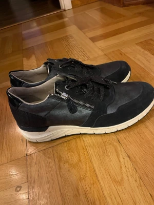 Svarta sneakers från Jana med dragkedja - Säljer ett par svarta sneakers från Jana med snygga detaljer som dragkedja på sidan och svarta glittriga skosnören. Skorna har en vit platt sula och är tillverkade i mocka och syntetmaterial. Rund tå och bekväm passform, perfekta för dig som gillar stilrena och coola sneakers.