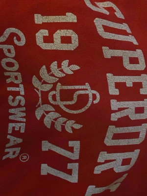 Röd Superdry hoodie med tryck - Snygg röd hoodie från Superdry med vit tryckt logga och text framtill. Tröjan har huva med vita snören, ribbade muddar och ficka på magen. Detaljer med vita ränder på ärmarna ger en sportig vibe. Mjuk insida i bomullsmix, perfekt för chill eller häng.