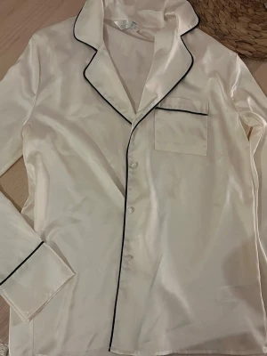 Vit satinblus med svarta detaljer - Säljer en vit långärmad blus i satin med svarta kanter och knappar framtill. Passar mer XS-S. Har en liten fläck men de syns inte så mycket