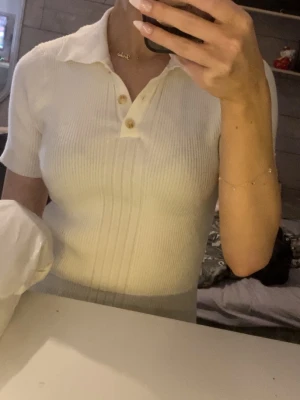 Vit ribbad polotopp med knappar - Säljer en vit ribbad polotopp med korta ärmar och snygg krage. Toppen har tre knappar framtill och är figurnära i modellen