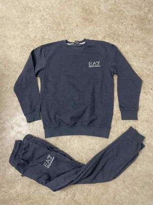 Mörkgrå mjukisbyxor från EA7 - Säljer ett par mörkgrå mjukisbyxor från EA7 Emporio Armani med logga tryckt på benet. Byxorna har elastisk midja med dragsko och ribbade muddar vid benslut. Perfekta för chill eller träning, tillverkade i mjukt bomullsmaterial.