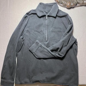 Svart overshirt från COS - Säljer en stilren svart overshirt från COS med två stora bröstfickor och en halv dragkedja framtill. Skjortan har klassisk krage och långa ärmar, tillverkad i mjuk bomull som ger en chill och avslappnad vibe.