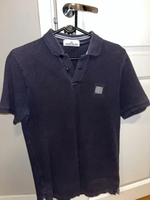 Svart kortärmad polotopp från Stone Island - Säljer en svart polotopp från Stone Island med klassisk krage och korta ärmar. Toppen har knappar vid halsen och en broderad logotyp på bröstet. Materialet är mjuk bomull och passformen är normal. Perfekt för dig som gillar stilrena och enkla plagg. 