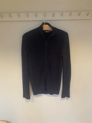 Svart ribbad kofta med dragkedja - Svart ribbstickad kofta från Dressmann med hel dragkedja framtill och ståkrage. Tröjan är långärmad och har en clean, stilren look som passar till många outfits. Materialet är mjukt och stickat, perfekt för kyligare dagar. Storleken är INTE S som det står. Den passar L då den är för stor i storlek.